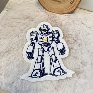 Könnte beinhalten: Ein Bügelbild aus weißem Filz mit einem Cartoon-Roboter mit blauer Umrandung. Der Roboter hat ein gelbes Gesicht mit einem einzigen Auge und einen gelben Stern auf der Brust.