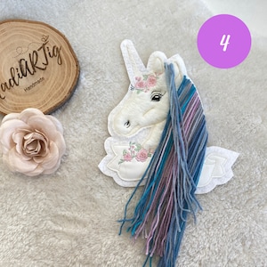 Könnte beinhalten: Weißer Filz-Einhorn-Applikationen mit einer rosa Blumenkrone und einer langen blauen, rosa und grauen Garnmähne. Das Einhorn befindet sich auf einem weißen Hintergrund mit einem Holzschild, auf dem "Craft Jig Handmade" steht, und einer rosa Blume.