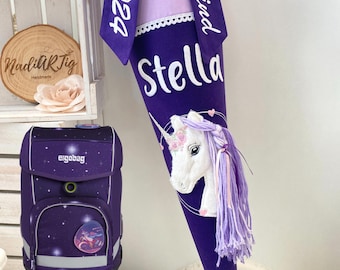 Könnte beinhalten: Lilafarbener Schulranzen mit Galaxie-Druck und Einhorn-Motiv. Ein lilafarbener und weißer Schultüte mit einem Einhorn-Applique und dem Namen "Stella" bestickt. Die Schultüte hat auch ein lilafarbenes Band mit dem Text "Schulkind 2024" darauf.