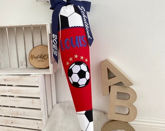 May include: A red and white soccer-themed school cone with the name "LOUIS" embroidered in blue. The cone features a soccer ball graphic, stars, and the year "2023" on a blue ribbon. The top is made of soccer ball patterned fabric.