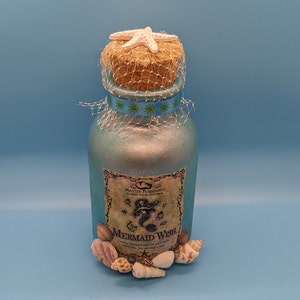 May include: A blue glass bottle with a cork stopper and a starfish on top. The bottle has a label that says "Mermaid Wish" and has a mermaid design. The bottle is decorated with seashells.