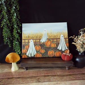Könnte beinhalten: Ein Halloween-Gemälde zeigt weiße Geister in einem Kürbisfeld mit einem Holzzaun. Die Kürbisse sind orange und der Himmel ist hellblau. Ein kleiner, roter, gehäkelter Kürbis und ein dekorativer Pilz sind ebenfalls vorhanden.
