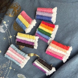 May include: A collection of eight small woven flags, each representing a different pride flag. The flags are made of colorful yarn and have a fringe on the bottom. The flags are arranged in a semi-circle on a blue fabric background.