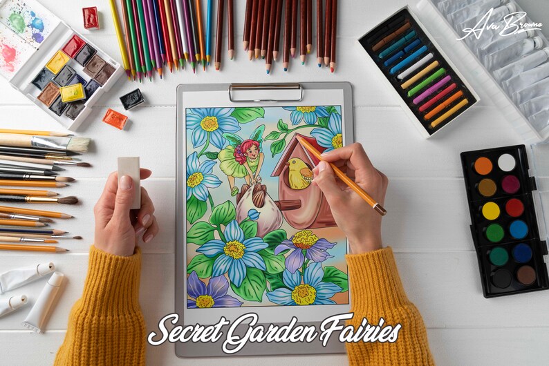 K&ouml;nnte beinhalten: Eine Person koloriert eine detaillierte Illustration einer Fee in einem Garten mit einem gelben Vogel in einem Vogelhaus. Die Illustration befindet sich auf einer Klemmbrett mit dem Text "Secret Garden Fairies". Die Person verwendet farbige Bleistifte und hat eine Vielzahl von Kunstmaterialien auf dem Tisch, darunter Wasserfarben, Pinsel und einen Radiergummi.