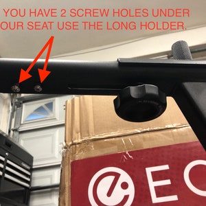 May include: Close-up of a black metal exercise equipment part with two screw holes. The text "IF YOU HAVE 2 SCREW HOLES UNDER YOUR SEAT USE THE LONG HOLDER." is visible in the image.