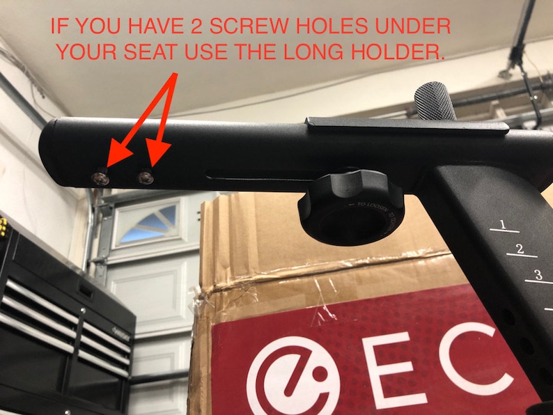 May include: Close-up of a black metal exercise equipment part with two screw holes. The text "IF YOU HAVE 2 SCREW HOLES UNDER YOUR SEAT USE THE LONG HOLDER." is visible in the image.