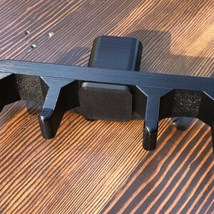 May include: Black plastic 3D printed tool with four prongs and a square base. The tool is designed to hold and secure objects.