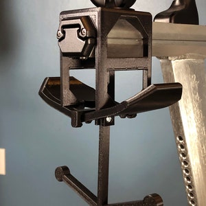 May include: A black, 3D-printed device with a complex design. It features a square frame with a knob on top, angled shelves, and a curved arm ending in two rounded supports. The device is mounted on a silver metal structure.