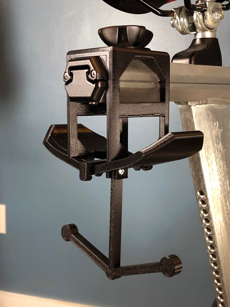 May include: A black, 3D-printed device with a complex design. It features a square frame with a knob on top, angled shelves, and a curved arm ending in two rounded supports. The device is mounted on a silver metal structure.