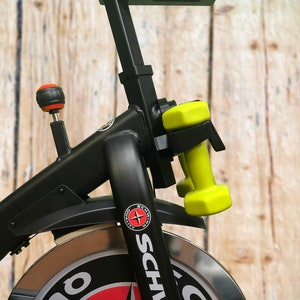 May include: Black exercise bike with a silver wheel and a black and red logo that says "Schwinn". There are two yellow dumbbells attached to the bike.