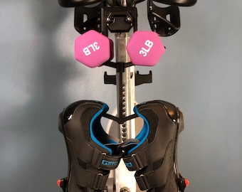 May include: Stationary exercise bike with black handlebars, a red and black seat, and two pink 3-pound dumbbells. Black cycling shoes with blue accents are attached to the pedals. The bike is set against a light blue wall.