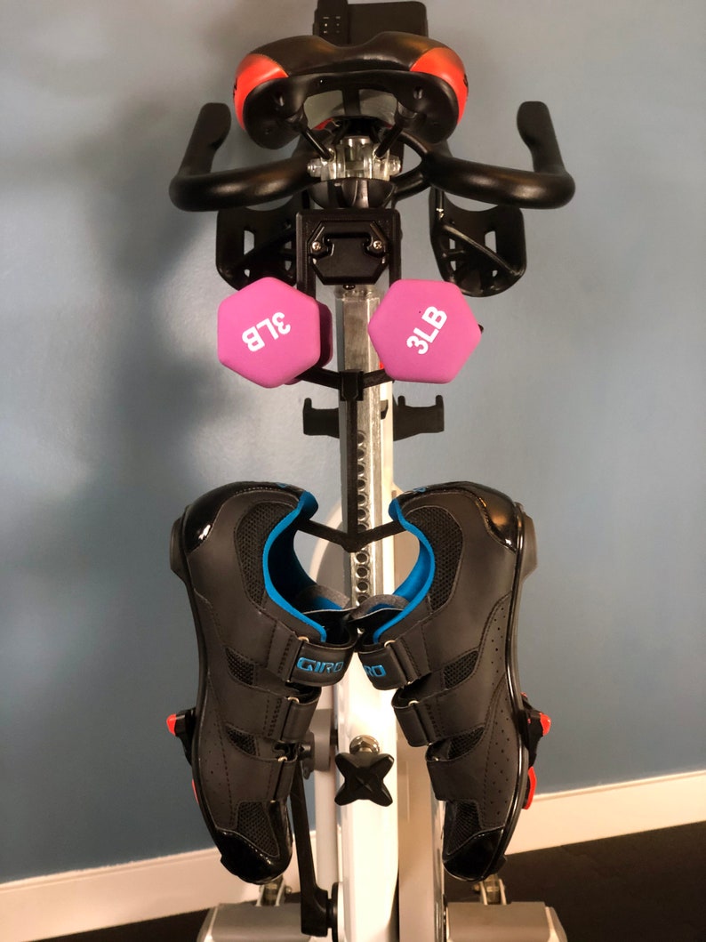 May include: Stationary exercise bike with black handlebars, a red and black seat, and two pink 3-pound dumbbells. Black cycling shoes with blue accents are attached to the pedals. The bike is set against a light blue wall.