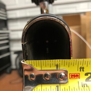 May include: A black bicycle seat post with a silver trim, measured with a yellow tape measure. The seat post opening is approximately 1 inch wide.