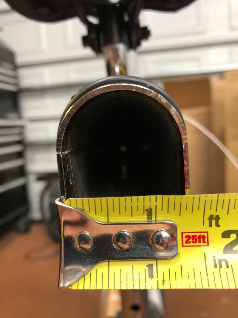 May include: A black bicycle seat post with a silver trim, measured with a yellow tape measure. The seat post opening is approximately 1 inch wide.