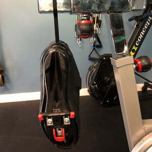 May include: A black cycling shoe with red cleats hanging from a silver exercise bike. The shoe is attached to a black strap.