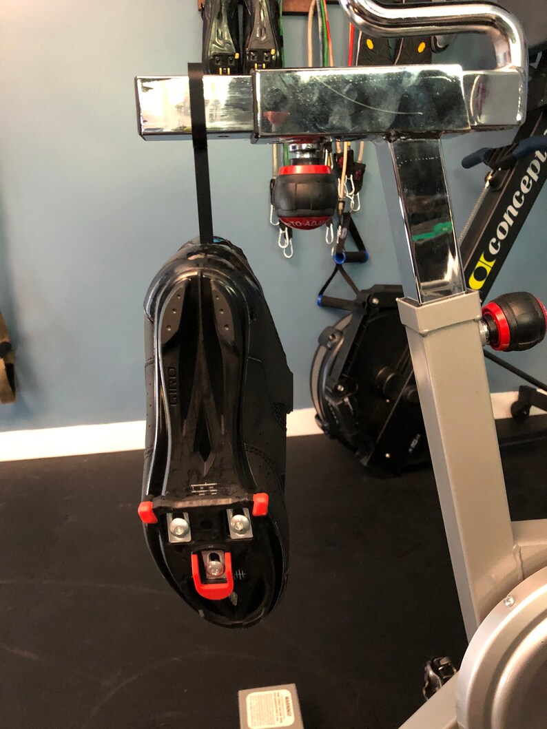 May include: A black cycling shoe with red cleats hanging from a silver exercise bike. The shoe is attached to a black strap.