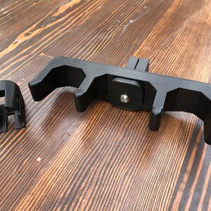 May include: Black 3D-printed device with a rectangular base and multiple curved arms, along with a separate cylindrical component. The device is designed to hold or support another object. The components are made of black plastic and sit on a wooden surface.