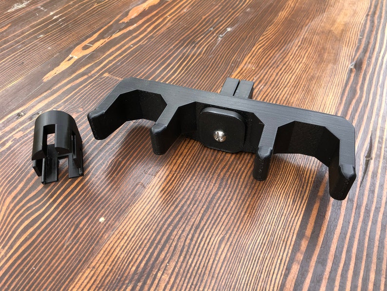 May include: Black 3D-printed device with a rectangular base and multiple curved arms, along with a separate cylindrical component. The device is designed to hold or support another object. The components are made of black plastic and sit on a wooden surface.