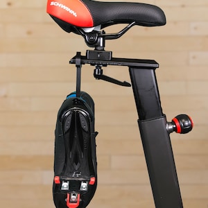 May include: A black exercise bike with a red and black seat that says "SCHWINN" on it. A black cycling shoe with red cleats is hanging from the bike.
