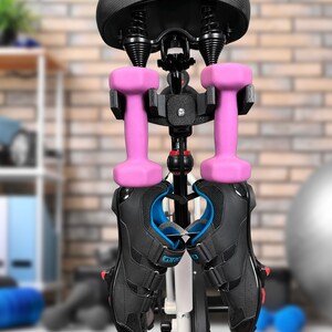 May include: A black bike seat with a white frame and a black and silver bike rack. Two pink dumbbells and two black cycling shoes are hanging from the rack.