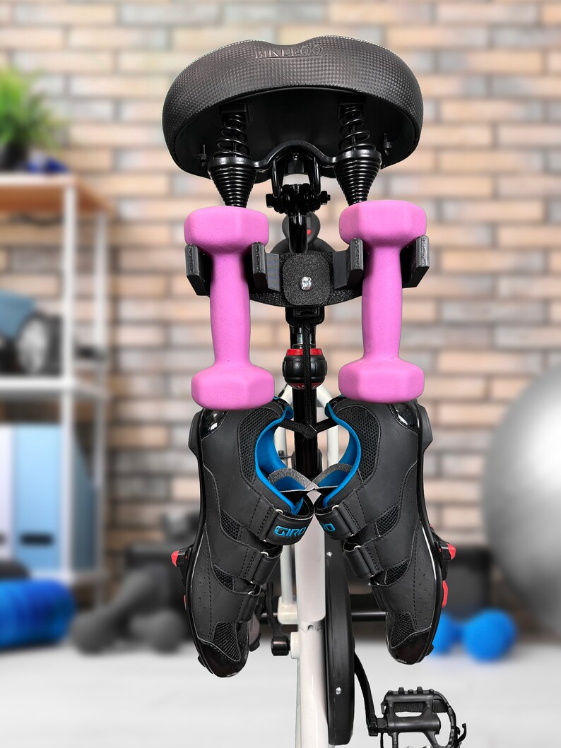May include: A black bike seat with a white frame and a black and silver bike rack. Two pink dumbbells and two black cycling shoes are hanging from the rack.