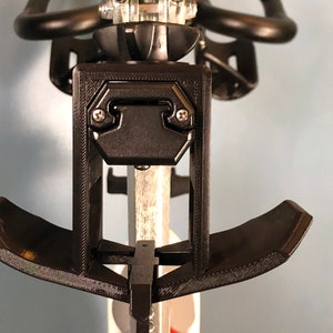 May include: Black 3D-printed bicycle accessory, featuring a rectangular bracket with two screws and a curved support. The design includes a central post and a curved, padded rest. The accessory is mounted on a bicycle seat post.