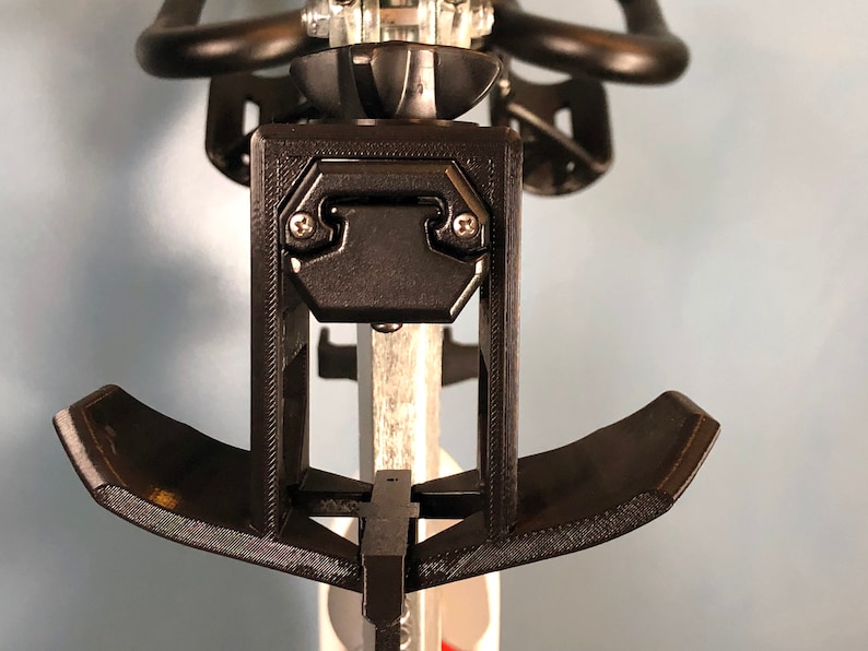 May include: Black 3D-printed bicycle accessory, featuring a rectangular bracket with two screws and a curved support. The design includes a central post and a curved, padded rest. The accessory is mounted on a bicycle seat post.