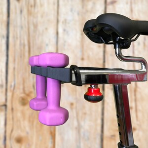 May include: A stationary exercise bike with a black seat and chrome frame. Two pink dumbbells are held in a black holder attached to the bike. The background is a light brown wooden wall.