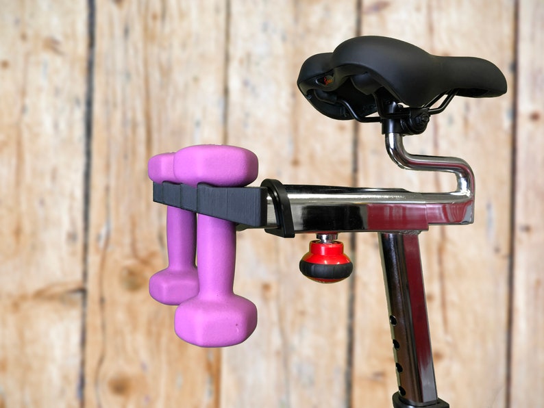 May include: A stationary exercise bike with a black seat and chrome frame. Two pink dumbbells are held in a black holder attached to the bike. The background is a light brown wooden wall.