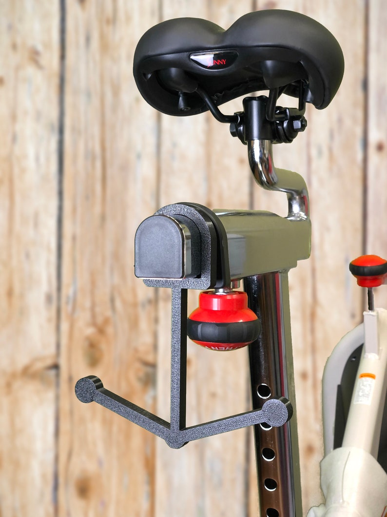May include: Stationary exercise bike with a black seat and a red and black resistance knob. The bike has a silver frame and a black support structure. The background is a light brown wooden wall.