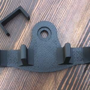 May include: Black plastic wall mount with three hooks and a circular hole. The mount is designed to hold items like a broom, mop, or rake.