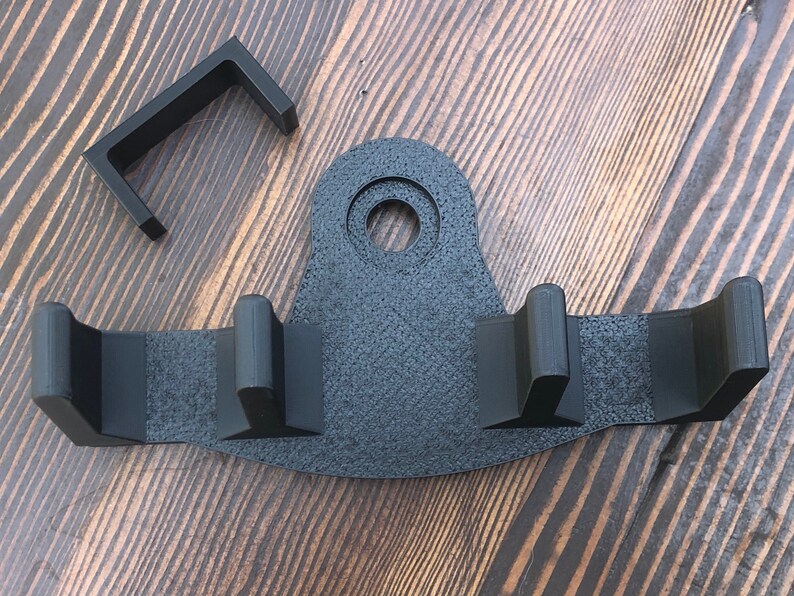 May include: Black plastic wall mount with three hooks and a circular hole. The mount is designed to hold items like a broom, mop, or rake.