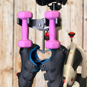 May include: An exercise bike with a black seat, a black metal dumbbell holder, two pink dumbbells, and a pair of black cycling shoes with blue accents. The bike has a wooden background.