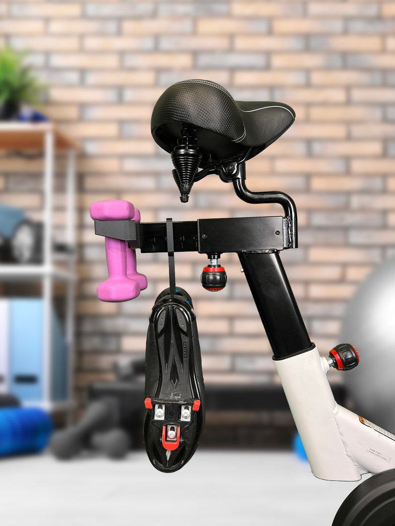 May include: A black exercise bike with a black seat and a white frame. A pair of pink dumbbells and a black cycling shoe are hanging from the bike.