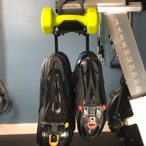 May include: A pair of black cycling shoes with cleats hanging from a black and yellow dumbbell holder. The shoes are suspended from a metal bar, with a scale marked from 2 to 20.