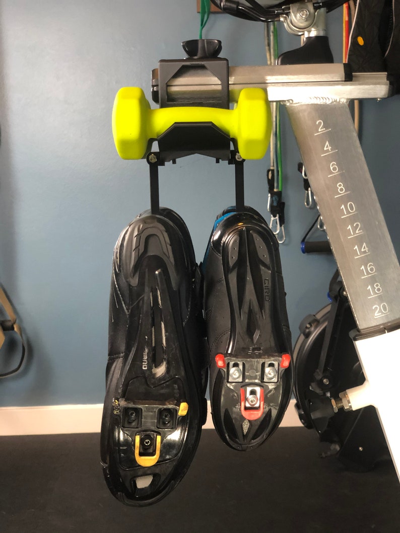 May include: A pair of black cycling shoes with cleats hanging from a black and yellow dumbbell holder. The shoes are suspended from a metal bar, with a scale marked from 2 to 20.