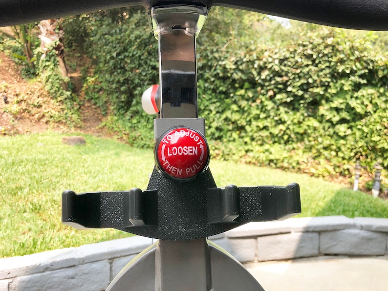May include: A red button with white text that reads "To Adjust Loosen Then Pull" on a silver exercise bike.