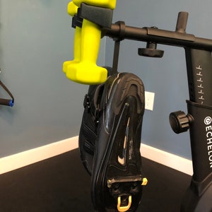 May include: A black exercise bike with a yellow dumbbell hanging from the handlebars. The bike has a digital display and a black seat. The dumbbell is hanging from a black hook.