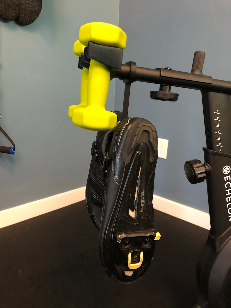 May include: A black exercise bike with a yellow dumbbell hanging from the handlebars. The bike has a digital display and a black seat. The dumbbell is hanging from a black hook.