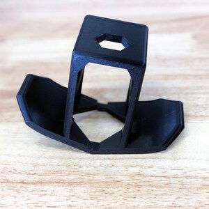 May include: A black 3D-printed phone stand with a hexagonal opening at the top. The stand has a rectangular top and two angled supports that connect to a base with curved edges. The stand is made of plastic.