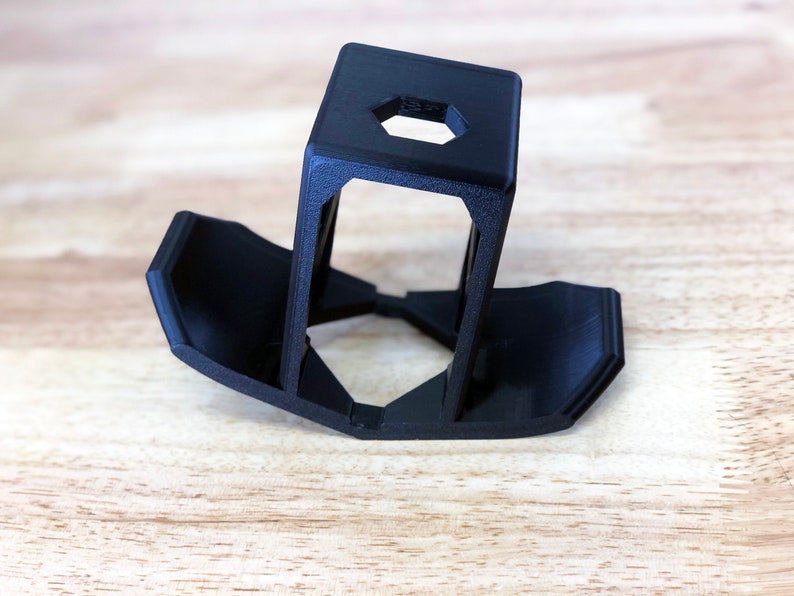 May include: A black 3D-printed phone stand with a hexagonal opening at the top. The stand has a rectangular top and two angled supports that connect to a base with curved edges. The stand is made of plastic.