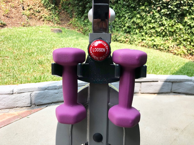 May include: Two purple dumbbells are attached to a black holder on a silver exercise machine. The machine has a red button that says "To Adjust Loosen Then Pull".