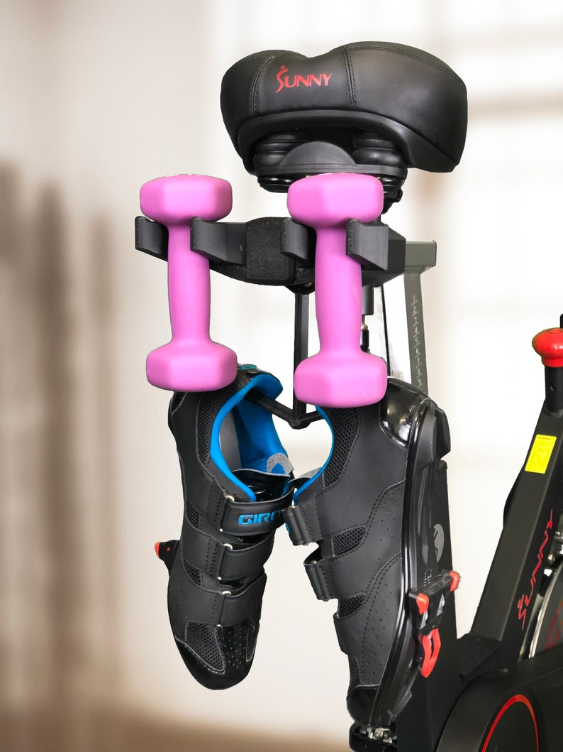 Pu&ograve; includere: Una cyclette nera con una sella nera su cui &egrave; stampata la parola "SUNNY" in rosso. Due manubri rosa sono appesi alla cyclette e un paio di scarpe da ciclismo nere sono appese sotto i manubri.