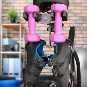 May include: A black exercise bike with a pink dumbbell holder attached to the seat post. Two pink dumbbells are hanging from the holder. A pair of black cycling shoes are hanging from the bike.