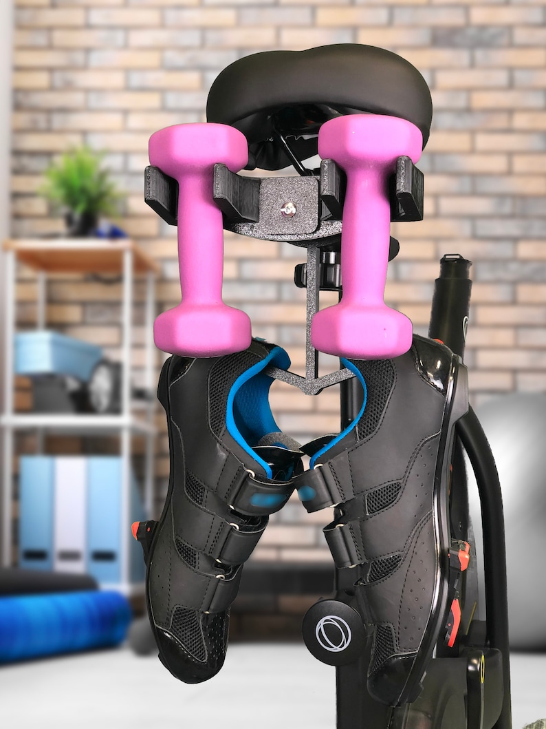 May include: A black exercise bike with a pink dumbbell holder attached to the seat post. Two pink dumbbells are hanging from the holder. A pair of black cycling shoes are hanging from the bike.