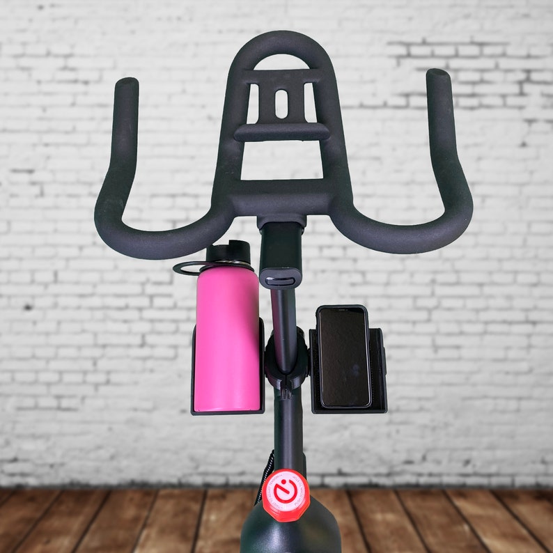May include: Black stationary exercise bike with a water bottle holder and a phone holder. The bike has a red button on the front.