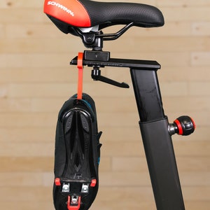 May include: A black exercise bike with a red and black saddle and a black shoe attached to the frame with a red strap.