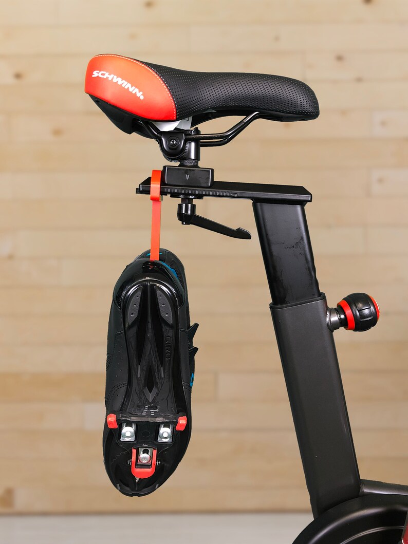 May include: A black exercise bike with a red and black saddle and a black shoe attached to the frame with a red strap.