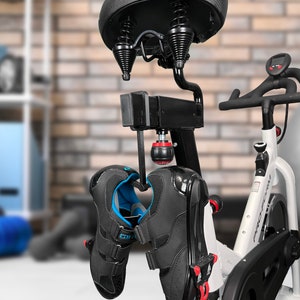 May include: Black and white cycling shoes hanging from a hook attached to a stationary bike. The bike has a black seat with springs and a white frame. The shoes have blue accents and a velcro strap.
