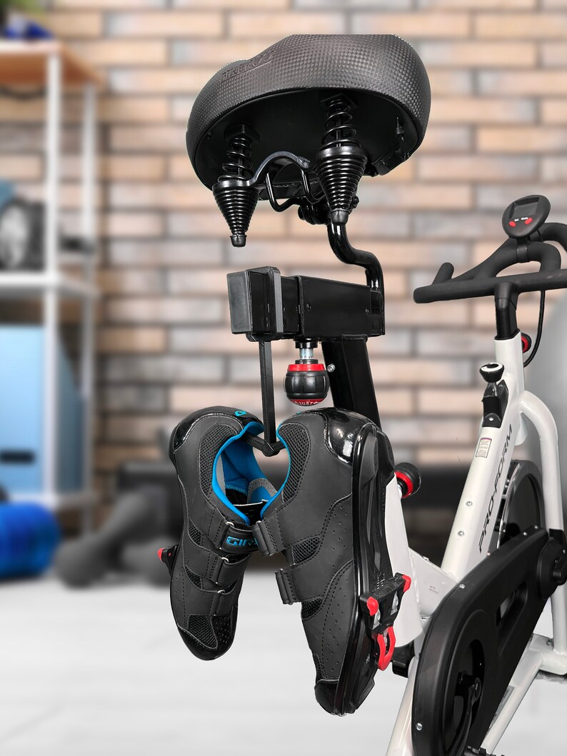 May include: Black and white cycling shoes hanging from a hook attached to a stationary bike. The bike has a black seat with springs and a white frame. The shoes have blue accents and a velcro strap.